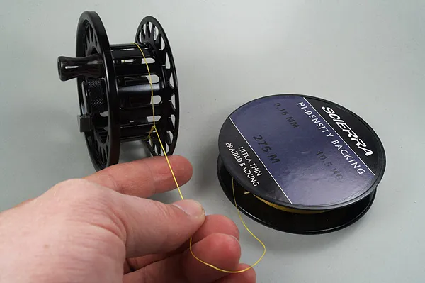 Backing attached - The backing is now attached to the spool. This is an open cage reel, which allows for the backing to pass into the cage after it has been attached.<br />
If the reel has a closed rim, you will have to pull the backing through the reel cage before attaching it to the spool, and make sure it's done through the right opening in the reel.<br />
On a ventilated reel like this one, you can also tie the backing to one of the bars in the axle.